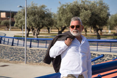 Mature, gray haired man with beard, sunglasses, white shirt and white headphones, dancing happily while listening to music, by a lake. Concept happiness, joy, music, disco, dancing, retirement.の写真素材
