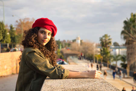 Portrait of a young and beautiful woman, brunette, with curly hair, with a green jacket and red beret, leaning on a stone wall. Concept beauty, fashion, trend, model, seduction.の写真素材