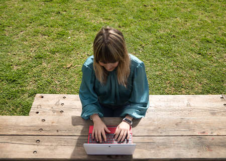 Young, pretty, blonde woman in green shirt, sitting on a wooden bench in a park, working with her laptop, view from above. Technology concept, tablet, notebook, teleworking, telecommuting, zenithal.の写真素材