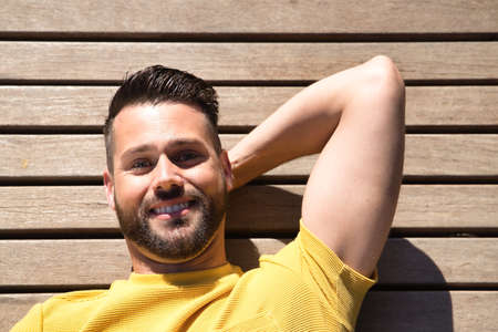 Portrait of handsome young man, with blue eyes and perfect smile, wearing yellow t-shirt, lying on a wooden slatted surface smiling happily. Concept beauty, fashion, happiness, trend.の写真素材