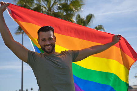 Young and handsome gay man, with a beard and green shirt, with blue eyes, perfect smile and waving a gay pride flag, smiling. Concept of gay pride, homosexual, lgtbi, pride day.の写真素材