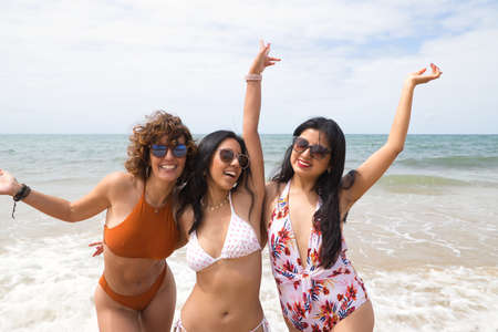 Attractive mature woman next to two young, pretty, brunette South American women in bikini and sunglasses, posing clutching a surfboard in fun attitude. Concept vacation, friends, multiethnic, summer.の写真素材