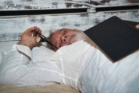 Mature, gray-haired, bearded man with a pair of glasses in his hand, in his pajamas, lying in bed asleep with a book on top of him. Concept reading, loneliness, retirement, pensioner, sleep, sleepingの写真素材