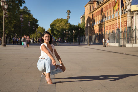 South American woman, young and beautiful, brunette, with white shirt and jeans, crouching and posing happily looking at the camera. Concept beauty, fashion, trend, model.の写真素材