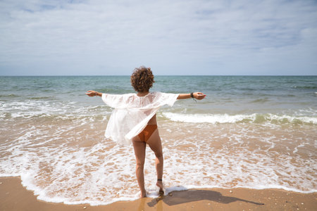 Attractive mature woman with curly hair and bikini, with open arms grateful and happy on the beach. Concept sea, sand, sun, beach, vacation, summer.の写真素材
