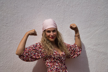 Middle-aged blonde woman, wearing a flower dress and a pink scarf of the fight against cancer, with arms up pulling muscle on a white background. Concept fight, disease, overcoming, cancer, breast.の写真素材