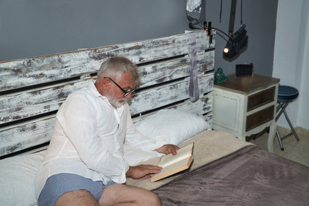 Mature, gray-haired man with beard and glasses, in pajamas, lying in bed reading a book quietly. Concept reading, loneliness, retirement, pensioner.の写真素材