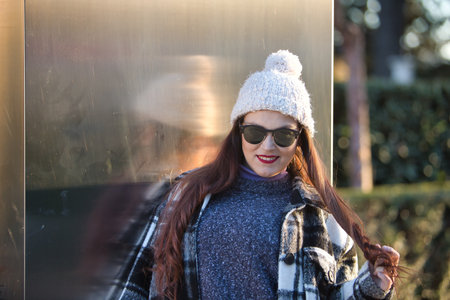 Young woman, beautiful, straight brown hair, sweater, coat, wool hat and sunglasses, leaning on a metal wall, playing with her hair, flirtatious and sensual. Concept beauty, autumn, winter, cold.の写真素材