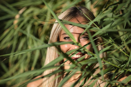 Portrait of young, beautiful, blonde woman with green eyes, looking smiling and happy through the green leaves of a plant. Concept beauty, eyes, nature, nature, looks, green, happiness.の写真素材