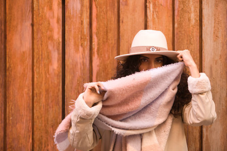 Young, pretty, brunette woman with curly hair and coat, scarf and hat, looking at camera while playing funny with her scarf, on wooden background. Concept beauty, fashion, autumn, winter, cold.の写真素材