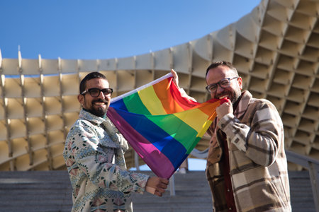Real marriage of gay couple, looking at camera while holding a gay pride flag as one of them bites it, happy and complicit. Concept lgtb, lgtbiq+, couples, in love, pride, flag.の写真素材
