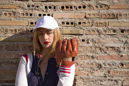 Portrait of young, beautiful, blonde woman with white cap, jacket, baseball glove and ball, leaning against a broken brick wall in the background. Concept beauty, sport, baseball, ball, glove.の写真素材