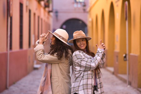 Two women, young and beautiful, with hats and coats, with their backs together and making the shape of a gun with their hands, happy and funny. Concept autumn, winter, cold, friendship, couples.の写真素材