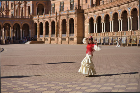 Young black and South American woman in a beige gypsy flamenco suit and red shawl, dancing in a beautiful square in the city of Seville in Spain. Concept dance, folklore, flamenco, art.の写真素材