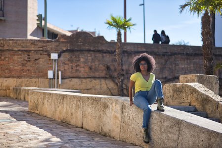 Young, beautiful, black woman with afro hair, wearing yellow t-shirt, jeans and sunglasses, sitting on a stone wall, relaxed, calm and sunbathing. Vacation concept, travel, current, modern.の写真素材