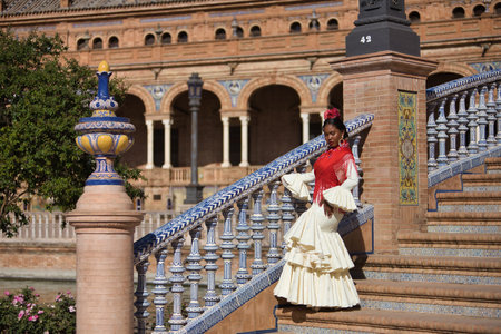 Young black and South American woman in a beige gypsy flamenco suit and red shawl, dancing in a beautiful square in the city of Seville in Spain. Concept dance, folklore, flamenco, art.の写真素材