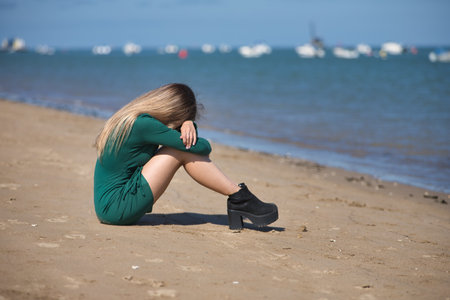 Young, beautiful, blonde woman, wearing a green dress, sitting on the shore of the beach, with her head between her knees and clutching her legs, crying, sad and depressed. Concept depression, sadnessの写真素材