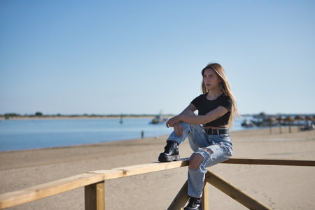 Young, pretty, blonde woman, wearing a black top, ripped jeans and tattoos, sitting on a railing, looking at infinity with the sea in the background. Concept of peace, tranquility, relaxation.の写真素材