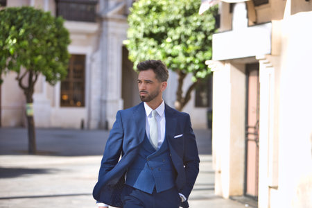 Young and attractive businessman, with beard and suit, walking along a city street with his hand in his pocket. Concept beauty, fashion, success, achiever, trend.の写真素材
