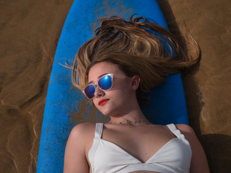 Portrait of young woman, blonde and beautiful, with a white bikini and sunglasses, lying on a blue surfboard, posing by the shore, with eighties style, seen from above. Beach concept, summer, eightiesの写真素材