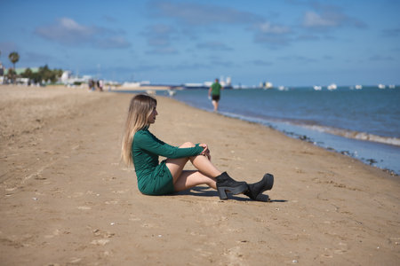 Young, beautiful, blonde woman in a green dress, sitting on the shore of the beach looking at the sea, alone, sad and depressed. Concept loneliness, depression, sadness.の写真素材
