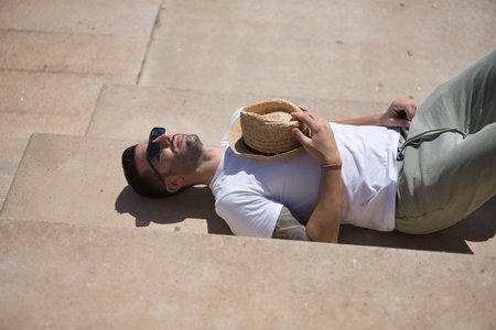 Young, attractive, Hispanic man wearing a white T-shirt, sunglasses and hat on his chest, lying on the floor sunbathing. Concept, vacation, relax, sun, snap, sleep.の写真素材