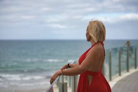 Mature, beautiful, blonde woman in a red dress, leaning on a glass railing looking at the sea in solitude and nostalgia. Concept beauty, fashion, trend, travel, maturity.の写真素材