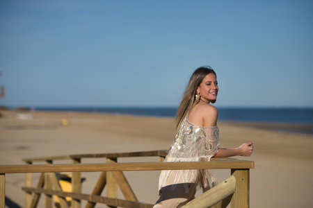 Young and beautiful woman with sequined and fringed shirt, leaning on a wooden railing, looking at the sea smiling and happy. Concept beauty, fashion, trend, empowerment.の写真素材