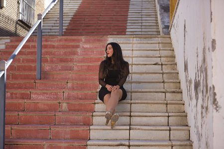 Young, beautiful, brunette woman in an elegant black dress, with a distracted look sitting on the stairs of a beautiful white Andalusian village. Concept beauty, fashion, trend, travel, rural.の写真素材
