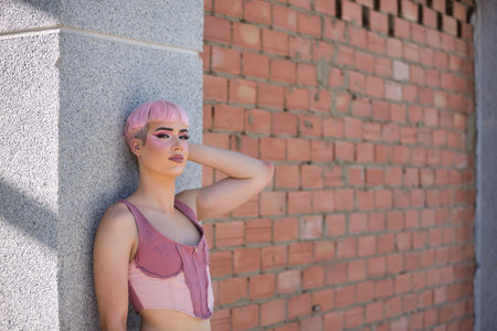 Young, attractive, gay man, heavily makeup, with pink hair, top and jeans, leaning against a gray wall looking at camera, relaxed and calm. Concept lgtbiq+, gay, pride, makeup, fashion, trend.の写真素材
