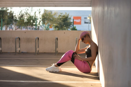 Young attractive gay man, heavily makeup, with pink hair, sunglasses, top and leather pants, alone, depressed and sad, sitting on the ground getting a sunbeam at sunset. LGTBIQ+ concept, gay, pride.の写真素材
