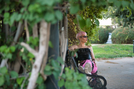 Young attractive gay man, heavily makeup, with pink hair, sunglasses, leather top and pants, relaxed and calm, sitting on park bench. LGTBIQ+ concept, gay, pride, makeup, fashion, trend.の写真素材