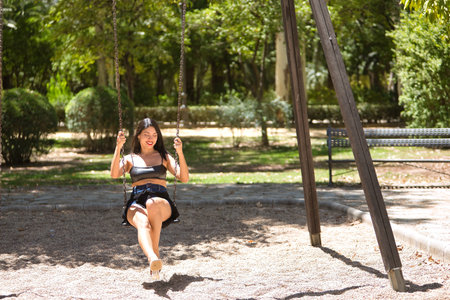 South American woman, young, pretty, brunette with leather top and short jeans, swinging on a swing very happy and smiling. Concept beauty, diversity, happiness, parks, games.の写真素材