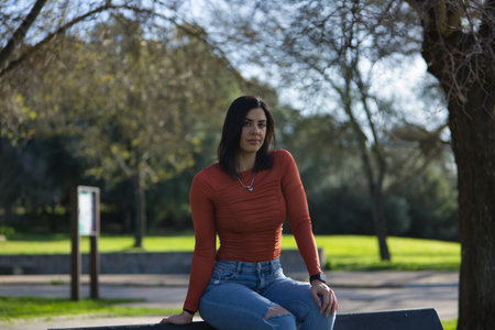 Young, pretty woman in orange t-shirt and jeans, looking at camera, relaxed and calm, sitting on a metal bench in a park. Fashion concept, beauty, tranquility, relaxation.の写真素材