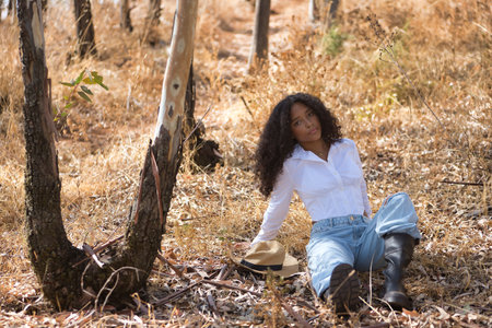 Latin woman, young, beautiful, brunette, with curly hair, wearing white shirt, jeans, boots and hat, sitting in a dry eucalyptus forest, relaxed and calm. Concept peace, relaxation, tranquility, forest.の写真素材
