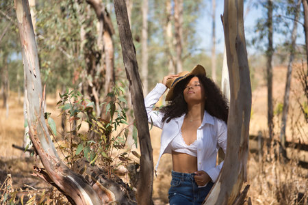 Latin woman, young, beautiful, dark-haired, with curly hair, with a white shirt and top, denim skirt and hat, closed eyes, among the trunks of a eucalyptus tree.の写真素材