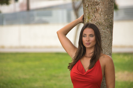 A young and beautiful woman, with green eyes, in an elegant red dress, looking at infinity leaning against the trunk of a tree. Concept beauty, fashion, trend, looks, nature.の写真素材