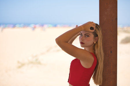 Young woman, beautiful, blue eyes, blonde, wearing red top, posing looking at camera, hands in her hair, leaning on a wooden pole with beach in background. Concept beauty, fashion, trend, femininity.の写真素材