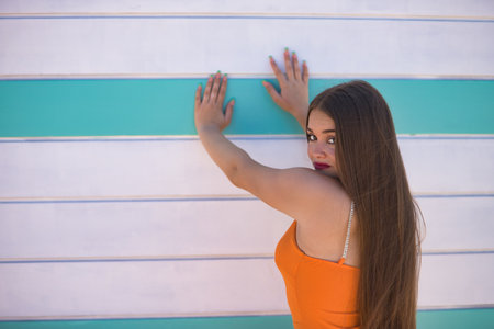 Portrait of beautiful young woman, with long brown hair, wearing an orange dress, with her back turned, looking over her shoulder, leaning against a beach hut.の写真素材