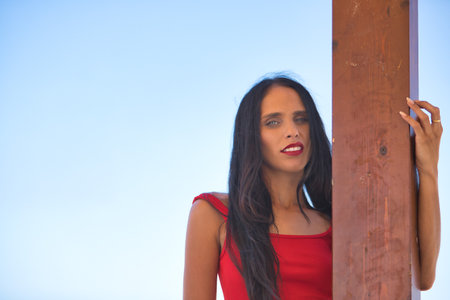 Portrait of young, beautiful, brunette, slim woman with a red top, posing looking at the camera, leaning on a wooden pole with the beach in the background. Concept beauty, femininity.の写真素材
