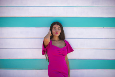 Young, beautiful, brunette woman with long hair, wearing a pink suit, with her hand on her head, smiling and flirtatious, leaning on a beach hut. Concept beauty, fashion, trendy, femininity.の写真素材