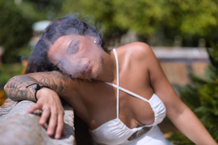 Young, dark-haired, beautiful, heavily tattooed woman in a white dress, sitting exhaling the white vapor smoke while vaping. Tattoos concept, nonconformism, rebellious, trend, fashion.の写真素材