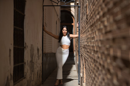 Young, beautiful, brunette woman in a white suit and heels, posing on a very narrow street leaning against the wall, receiving the sun's rays. Concept beauty, fashion, trend, city.の写真素材