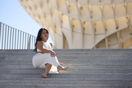 Young, beautiful, brunette, latina woman, wearing a white dress and heels, posing looking at camera, relaxed and calm sitting on stairs. Concept beauty, fashion, trend, city.の写真素材