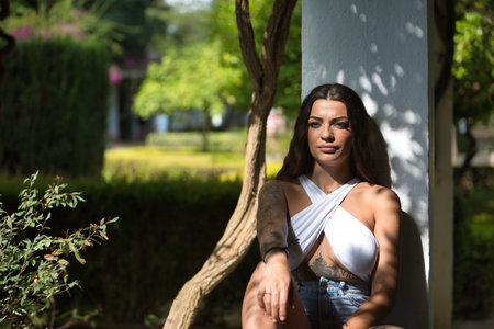 Young woman, brunette, beautiful, very tattooed, with white top and jeans, distracted and thoughtful, leaning on a column receiving the rays of the sun. Concept tattoos, nonconformism, rebel, trend.の写真素材
