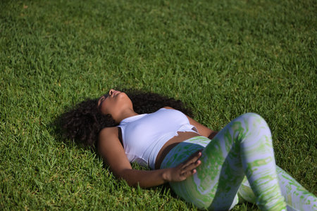 Latin woman, young, beautiful, brunette, with curly hair, wearing sports clothes, lying on the grass of a park resting after exercise. Concept sport, exercise, rest, active.の写真素材