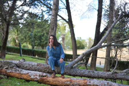Young, beautiful, brunette, Hispanic woman, wearing a denim suit, distracted and dreamy, with her hand on her chin, sitting on the fallen trunk of a tree, in nature. Concept fashion, beauty, trendy.の写真素材