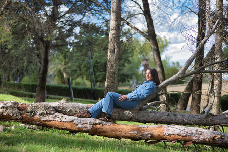 Young, beautiful, brunette, Hispanic woman, wearing a denim suit, smiling and happy, lying on the fallen trunk of a tree, in nature, receiving the sun's rays. Concept fashion, beauty, femininity.の写真素材