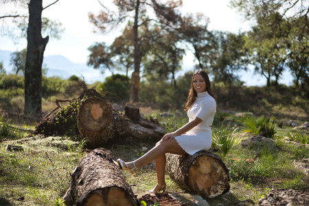 Pretty young woman in white sweater and skirt, smiling and happy looking at camera sitting on tree trunk in nature. Concept beauty, trend, femininity.の写真素材