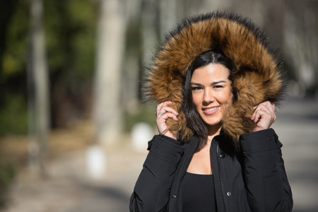 Portrait of a young, beautiful, dark-haired woman, looking at the camera, wrapping herself up with the fur collar of her coat, on a cold and sunny day. Concept winter, cold, autumn, weather.の写真素材